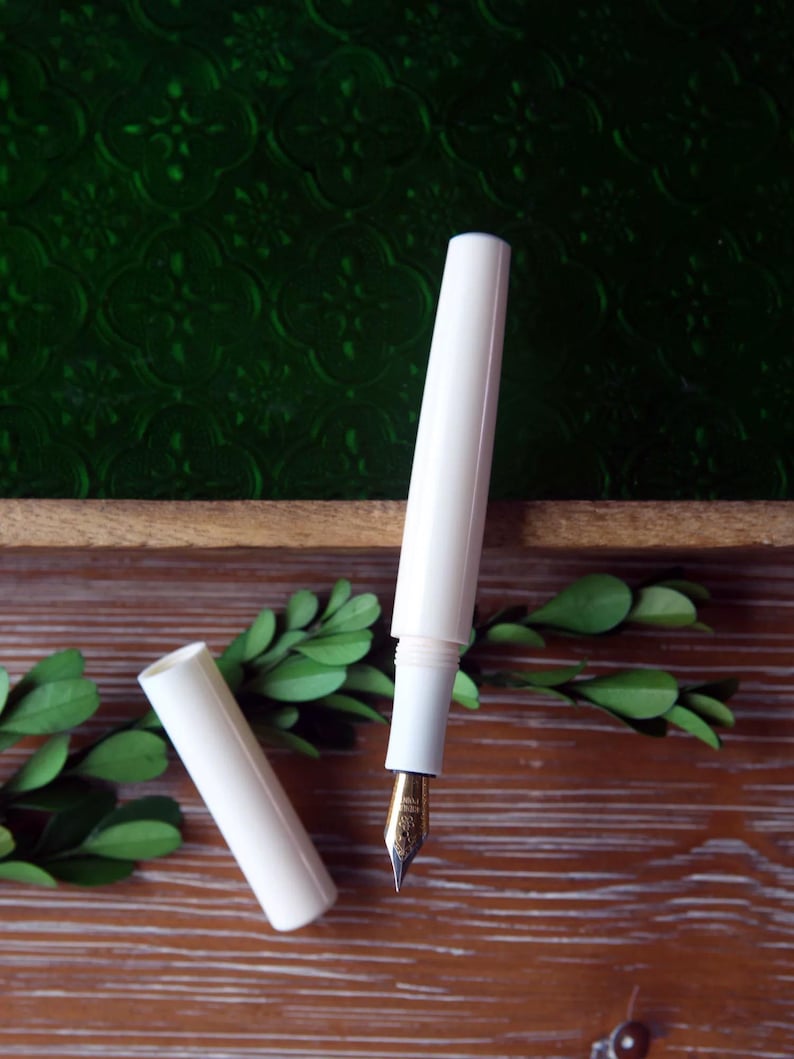 May include: A white fountain pen with a gold nib and a matching white cap. The pen is lying on a wooden surface with green foliage.