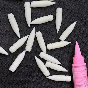 May include: A collection of white, bullet-shaped applicators scattered on a dark surface. A pink bottle with a pointed applicator tip is visible on the right side of the image. The applicators appear to be made of a soft, porous material.