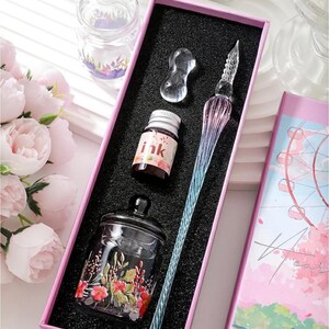 K&ouml;nnte beinhalten: Ein Kalligraphie-Set in einer rosa Schachtel mit einem Glas-Dip-Pen mit blauem und lila Farbverlauf, Tintenflaschen und einem kleinen Glas mit floralen Akzenten. Das Set enth&auml;lt ein klares Glas-Tintenfass und eine klare Glas-Stifthalterung.