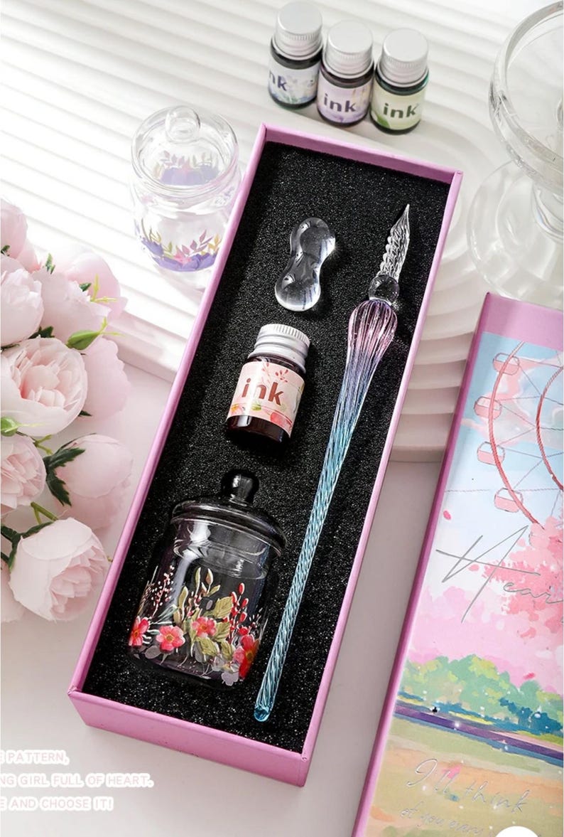 K&ouml;nnte beinhalten: Ein Kalligraphie-Set in einer rosa Schachtel mit einem Glas-Dip-Pen mit blauem und lila Farbverlauf, Tintenflaschen und einem kleinen Glas mit floralen Akzenten. Das Set enth&auml;lt ein klares Glas-Tintenfass und eine klare Glas-Stifthalterung.