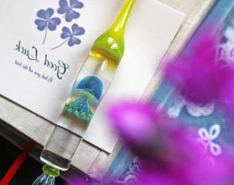 May include: A clear glass dip pen featuring a bright yellow handle and a blue jellyfish figure encased within the barrel. The pen has a pointed, fluted glass nib at the tip and rests on a white card printed with purple clover icons and the text Good Luck.