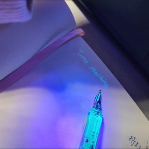 May include: A notebook with the words "Time Machine" written in light blue ink. A clear, turquoise pen is positioned on the page. The notebook has a purple ribbon bookmark.