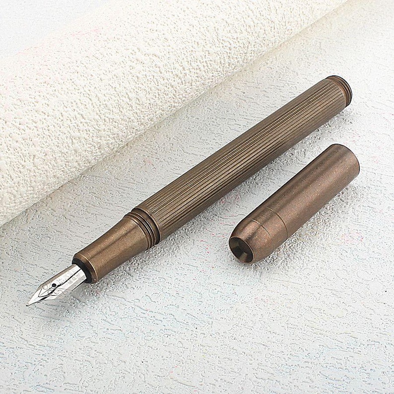 Puede incluir: Una pluma estilogr&aacute;fica de color bronce con un cuerpo acanalado y un plum&iacute;n plateado. El capuch&oacute;n est&aacute; separado y colocado junto a la pluma. La pluma est&aacute; sobre una superficie blanca texturizada. Un instrumento de escritura.