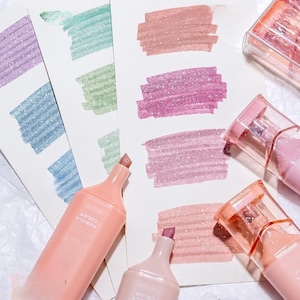 May include: A collection of pastel-colored highlighters with glittery ink, each with a different shade. The highlighters are peach-colored with the text "NEBULA COLOR" printed on them. Swatches of the ink are shown on white paper, including purple, blue, green, and pink.
