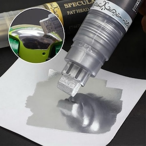 Puede incluir: Un rotulador metálico plateado con punta rectangular aplica pintura sobre una superficie blanca. El rotulador está etiquetado "SPECULA FAT HEAD" en letras doradas sobre un fondo negro. Un primer plano muestra la aplicación del rotulador sobre una superficie brillante y reflectante.