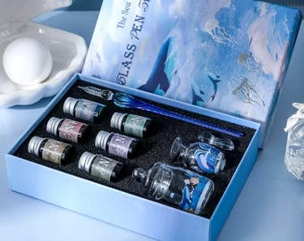 Op de afbeelding: Een blauwe geschenkdoos met het opschrift The Sea Land Glass Pen Set, bevattende een blauwe glazen dompelpen, zes kleine inktflesjes met zilveren doppen en twee doorzichtige glazen inktpotjes met blauw-witte illustraties van de oceaan.