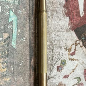 Puede incluir: Una pluma estilogr&aacute;fica dorada con un cuerpo estriado y un plum&iacute;n. La pluma se muestra verticalmente sobre un fondo estampado con motivos florales y figurativos. El dise&ntilde;o de la pluma sugiere un instrumento de escritura cl&aacute;sico.