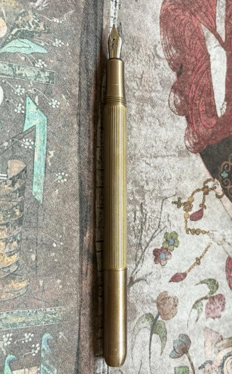 Puede incluir: Una pluma estilogr&aacute;fica dorada con un cuerpo estriado y un plum&iacute;n. La pluma se muestra verticalmente sobre un fondo estampado con motivos florales y figurativos. El dise&ntilde;o de la pluma sugiere un instrumento de escritura cl&aacute;sico.