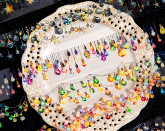 May include: A collection of small, colorful, translucent light bulb-shaped charms and stickers arranged on a decorative, cream-colored, perforated circular plate. The items feature vibrant, multi-colored patterns and star shapes.