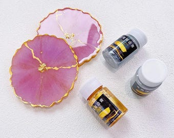 May include: Two round, scalloped-edge resin coasters in shades of pink with gold leaf accents and gold-painted rims, accompanied by three small plastic bottles containing gold and black metallic paint with white screw-on caps and black labels.