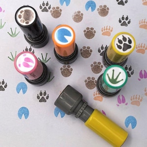 Peut inclure: Plusieurs tampons colorés avec des empreintes de pattes d'animaux, notamment des pattes de chien, de grenouille et de sabots de cheval. Les tampons sont disposés sur une surface blanche recouverte d'empreintes de pattes.