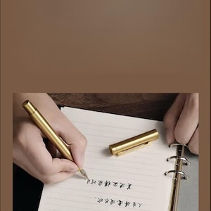 Puede incluir: Un bol&iacute;grafo dorado escribe sobre papel rayado. El bol&iacute;grafo est&aacute; junto a su capuch&oacute;n. El papel est&aacute; en un cuaderno con lomo dorado. El fondo es de color marr&oacute;n liso.