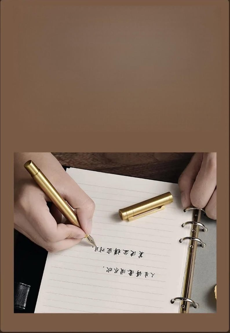 Puede incluir: Un bol&iacute;grafo dorado escribe sobre papel rayado. El bol&iacute;grafo est&aacute; junto a su capuch&oacute;n. El papel est&aacute; en un cuaderno con lomo dorado. El fondo es de color marr&oacute;n liso.