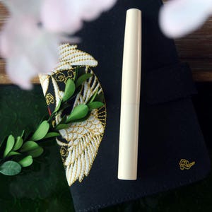 May include: A black notebook with a gold embroidered design of a bird with wings spread. A white pen is lying on top of the notebook.