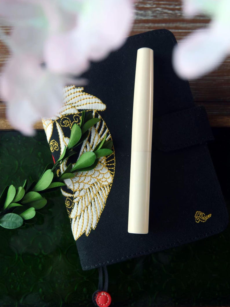 May include: A black notebook with a gold embroidered design of a bird with wings spread. A white pen is lying on top of the notebook.