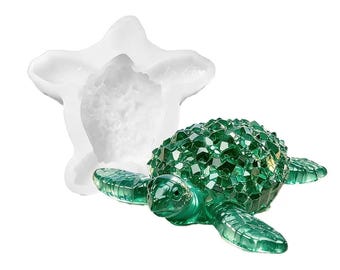 May include: A white silicone mold shaped like a sea turtle positioned behind a finished green sea turtle figurine with a faceted, crystal-like shell texture. The figurine has a translucent appearance and a detailed head, flippers, and body.