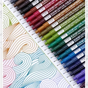 Puede incluir: Una colección de bolígrafos de punta fina de colores, dispuestos diagonalmente sobre una superficie blanca con un diseño. Los bolígrafos están etiquetados como "EXQUISITE GRAPHIC NEEDLE PEN" en varios colores, incluyendo negro, marrón, rojo, verde y azul. La obra presenta líneas onduladas.