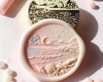 May include: A gold-toned metal wax seal stamp featuring an engraved night seascape with a crescent moon, stars, and waves. Below it, a pale pink wax seal impression displays the same detailed coastal scene with seashells and a starfish.