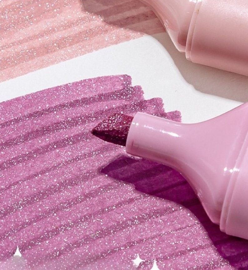 May include: Close-up of a pink highlighter with a glittery, dark pink tip, creating a vibrant, sparkling effect on a white surface. The highlighter is shown in use, with strokes of pink and light pink glittery ink.