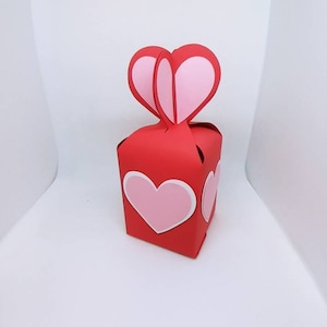 May include: A red paper gift box with a heart-shaped handle and pink heart accents. The box features a large pink heart on the front and side, outlined in white. The box is designed for Valentine's Day or other special occasions.