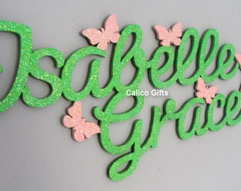 May include: Green glitter name sign with the names "Isabelle Grace" and a pink glitter star. The names are in a cursive font and there are pink glitter butterflies around the names.
