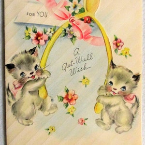 May include: A vintage greeting card featuring two fluffy gray kittens holding a large yellow wishbone. The card includes a pink ribbon, colorful floral accents, and the printed text FOR YOU and A Get-Well Wish against a diagonal striped background.