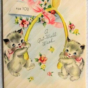 May include: A vintage greeting card featuring two fluffy gray kittens holding a large yellow wishbone. The card is decorated with pink ribbons and colorful flowers, with the text FOR YOU and A Get-Well Wish printed in black script on the front.
