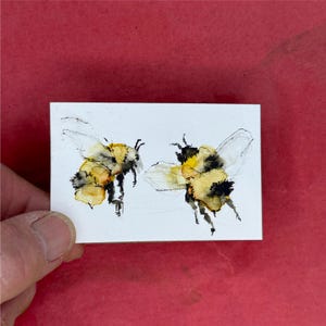 May include: A small rectangular white card featuring a watercolor illustration of two bumblebees. The bees are rendered in shades of yellow, black, and grey with delicate ink outlines, showing the insects in flight against a plain white background.