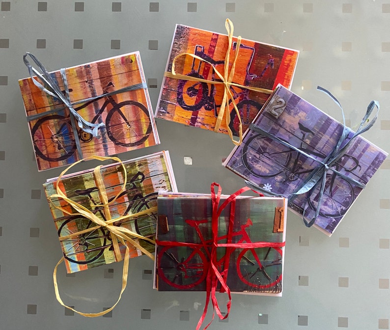 Puede incluir: Cinco tarjetas coloridas con ilustraciones de bicicletas. Cada tarjeta tiene un esquema de color y un dise&ntilde;o diferente. Las tarjetas est&aacute;n atadas con una cuerda.