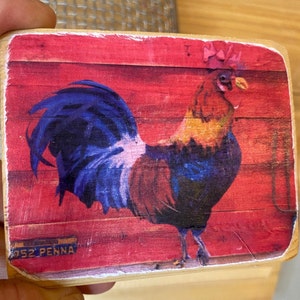 May include: A wooden sign with a red background and a painted image of a rooster with a pink bow on its head. The rooster is facing the right side of the sign. The sign has the text "952 PENNA" in the bottom left corner.