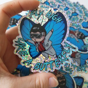 May include: A glittery sticker with a cartoon character dressed in a white suit and sunglasses, flying like a butterfly with blue wings. The sticker says "Fly High Like A Butterfly".