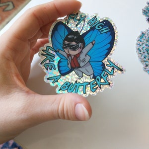 May include: A holographic sticker featuring a cartoon character wearing sunglasses and a white suit, flying like a butterfly. The sticker has the text "Fly High Like a Butterfly" in a blue and white font.