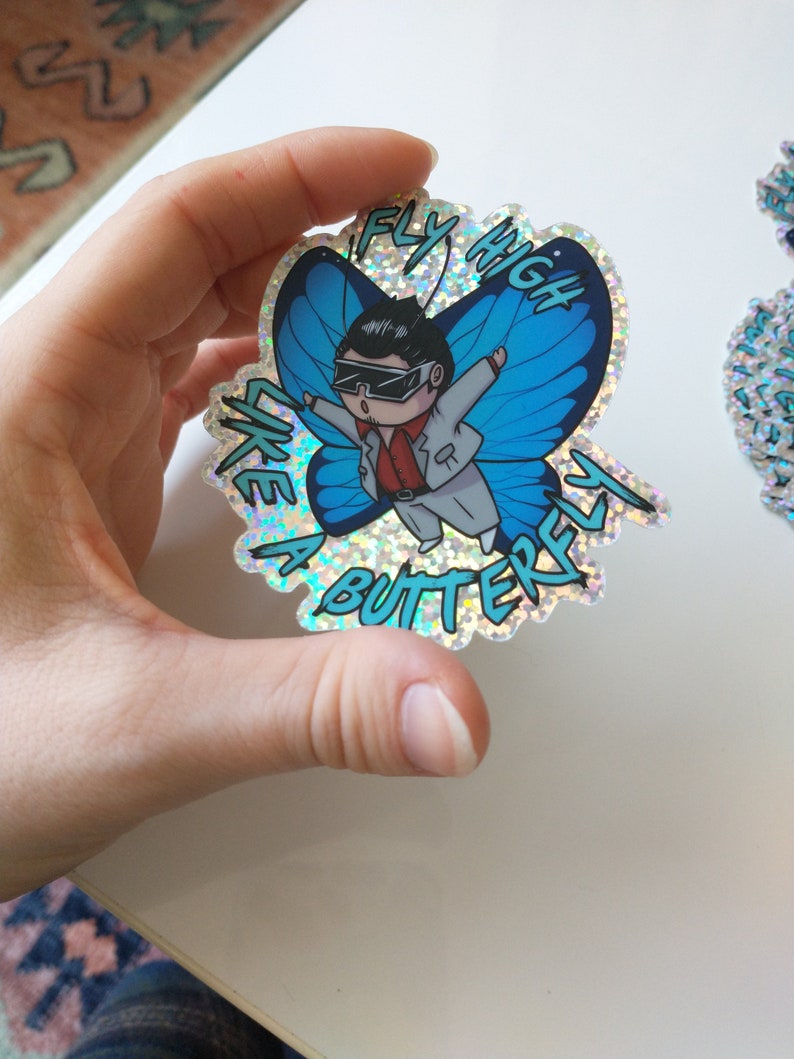 May include: A holographic sticker featuring a cartoon character wearing sunglasses and a white suit, flying like a butterfly. The sticker has the text "Fly High Like a Butterfly" in a blue and white font.