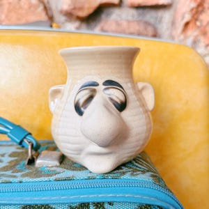 May include: A beige ceramic vase with a face design. The vase has a large nose, closed eyes, and small ears. The vase is sitting on a blue and green patterned surface, with a yellow background.