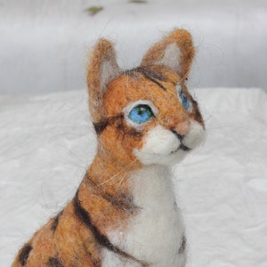 May include: A felted brown and white tabby cat with blue eyes. The cat is sitting on a white surface and looking to the right.