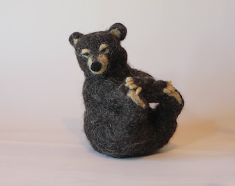 May include: A handmade, dark gray felt bear figurine with tan accents on its paws and ears. The bear is in a playful pose, with its legs curled up. The bear has a black nose and closed eyes.