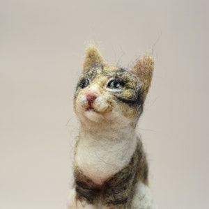 May include: A hand-felted cat figurine with brown, white, and black fur. The cat is sitting up with its head tilted up and its eyes open. The figurine is made of wool and is very detailed.