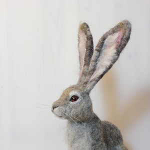 Puede incluir: Una escultura de conejo de fieltro de aguja gris y blanco con orejas largas y ojos rojos. El conejo está de pie sobre una superficie blanca.