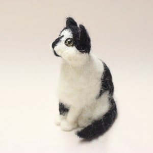 May include: A needle felted black and white cat figurine. The cat is sitting with its head tilted up and its eyes looking to the side. The cat is made of wool and has a realistic appearance.