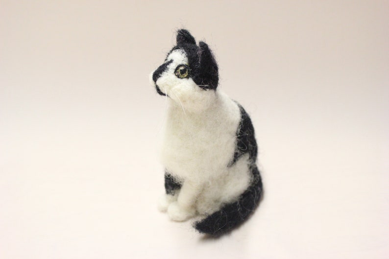 May include: A needle felted black and white cat figurine. The cat is sitting with its head tilted up and its eyes looking to the side. The cat is made of wool and has a realistic appearance.