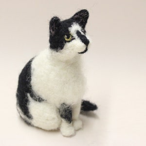 May include: A black and white needle felted cat figurine. The cat is sitting with its head tilted to the side and its eyes are open. The cat has a white chest and belly, and black markings on its face, back, and tail.