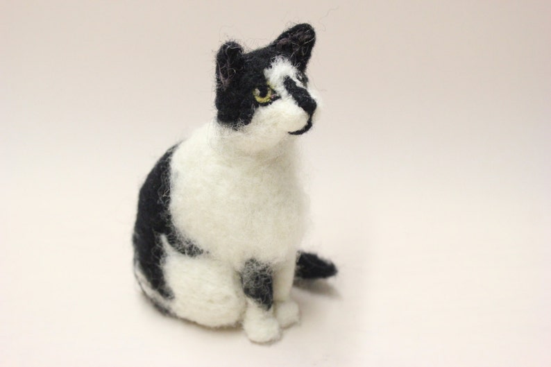 May include: A black and white needle felted cat figurine. The cat is sitting with its head tilted to the side and its eyes are open. The cat has a white chest and belly, and black markings on its face, back, and tail.