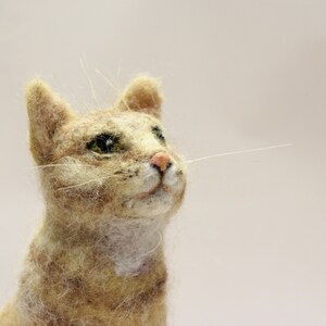 May include: A close-up of a needle felted cat sculpture. The cat is light brown with white markings and has green eyes. The cat is looking up and has a soft, fluffy texture.