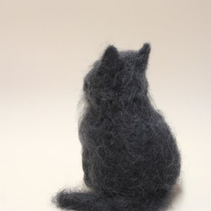 May include: A small, gray, felted cat figurine sitting on a white surface. The cat is facing away from the viewer, with its tail curled around its body.