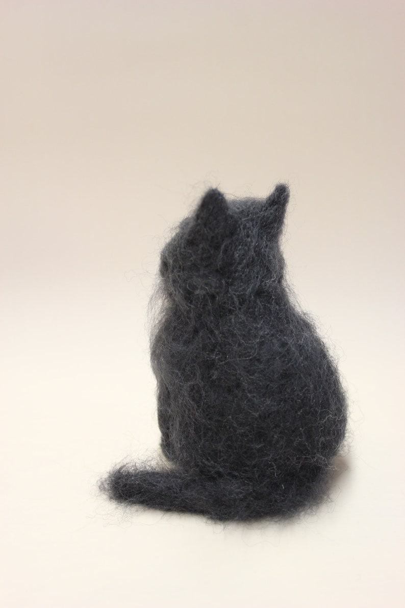 May include: A small, gray, felted cat figurine sitting on a white surface. The cat is facing away from the viewer, with its tail curled around its body.