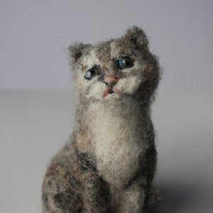May include: A gray and white needle felted cat figurine with blue eyes. The cat is sitting with its paws tucked underneath it.