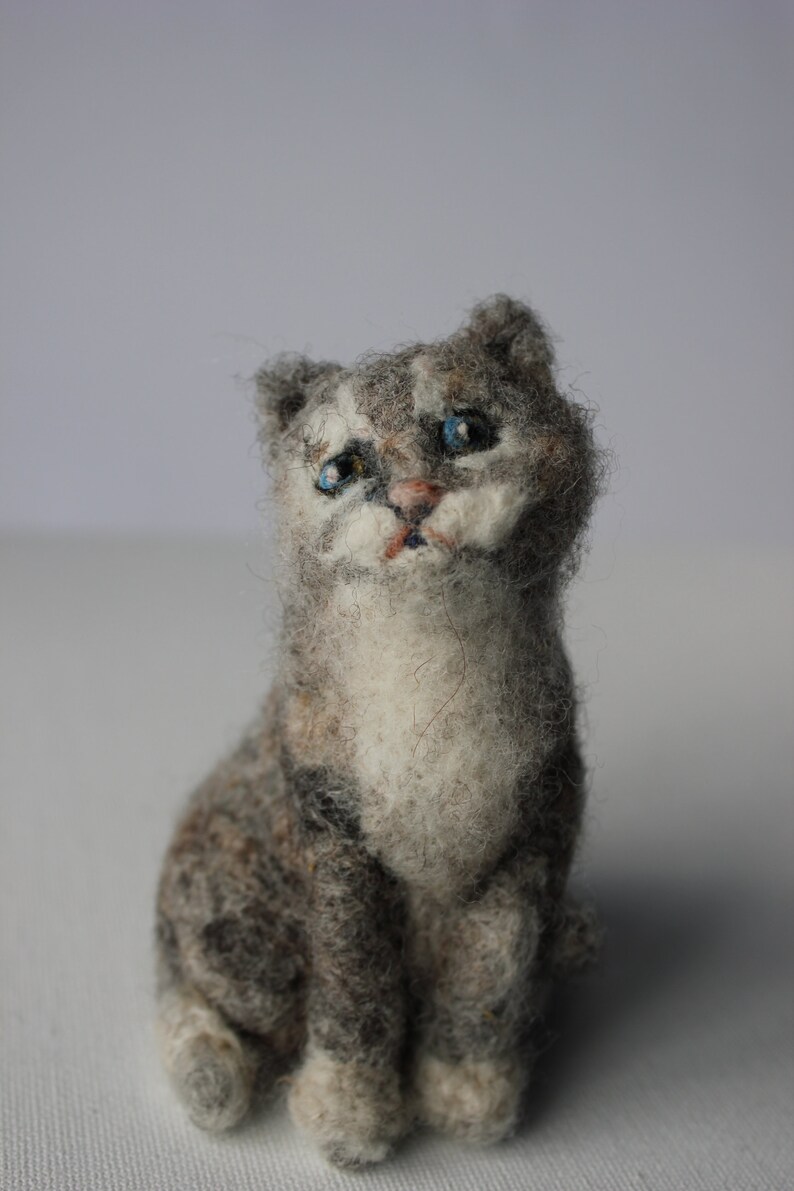 May include: A gray and white needle felted cat figurine with blue eyes. The cat is sitting with its paws tucked underneath it.