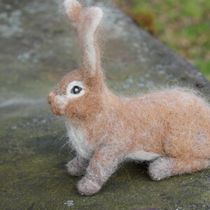 May include: A brown and white needle felted rabbit with long ears. The rabbit is sitting on a gray textured surface.