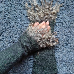 May include: A pair of dark forest green fingerless gloves featuring a textured, felted surface. The cuffs are adorned with long, curly, grey and silver wool locks that extend outward, creating a voluminous, shaggy appearance at the wrist.