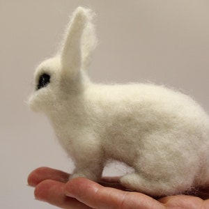 May include: A white, fluffy, needle-felted rabbit figurine with black eyes. The rabbit is sitting on a person's hand.
